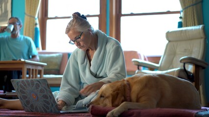 Mature woman in bathrobe working on laptop computer with dog next to her and man in background on phone.