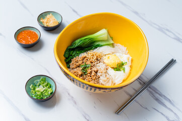 A vibrant yellow bowl of Asian noodles topped with minced pork, poached egg, and bok choy, served with green onions and chili sauce on a white marble surface, ideal for food photography