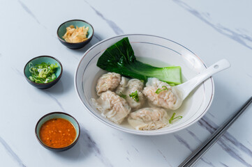 A delicate bowl of wonton soup with bok choy, served with fresh green onions, fried garlic, and spicy chili sauce on a white marble table, complemented by chopsticks