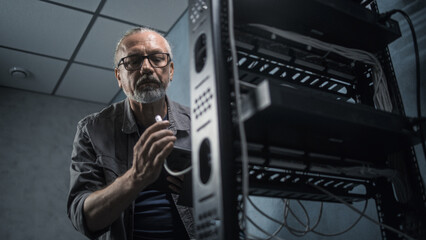 Mature engineer inserts the cable into the communications equipment, uses tablet computer. Male system administrator works in server room. Local area network or web server maintenance. Tech support.