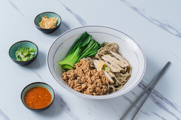 A white bowl of Asian noodles topped with minced pork, sliced chicken breast, and bok choy, served with green onions, fried garlic, and chili sauce on a marble surface, ideal for food photography