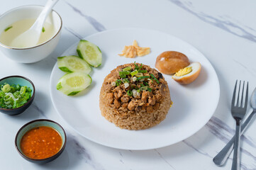 A white plate of Thai-style rice topped with minced chicken, served with sliced cucumber, a halved boiled egg, green onions, fried garlic, and chili sauce, alongside a soup bowl,