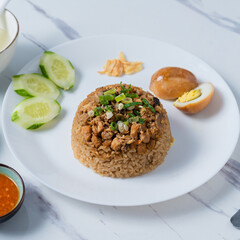 A white plate of Thai-style rice topped with minced chicken, served with sliced cucumber, a halved boiled egg, green onions, fried garlic, and chili sauce, alongside a soup bowl,