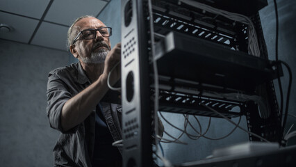 Mature engineer inserts the cable into the communications equipment, uses tablet computer. Male...
