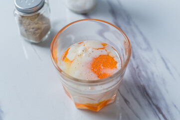 A glass of soft-boiled egg seasoned with salt and pepper, with a touch of soy sauce, placed on a marble surface, ideal for breakfast content or culinary presentations focused on nutritious meals.