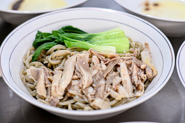 A close-up of a white bowl filled with Asian noodles, topped with sliced chicken and bok choy, set on a dark surface, perfect for showcasing traditional Asian cuisine in food photography