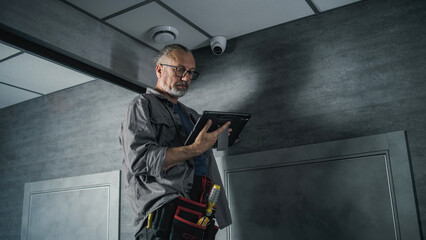 Male installer adjusts the angle of CCTV camera in office, sets up it using digital tablet computer with professional program. Monitoring and tracking. Security system, video surveillance and privacy.