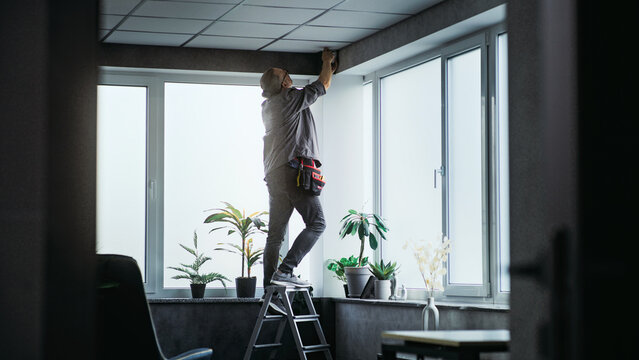Professional male installer in uniform stands on ladder and screws surveillance camera to the ceiling in office room using tools. Monitoring and tracking. CCTV system, security and privacy concept.