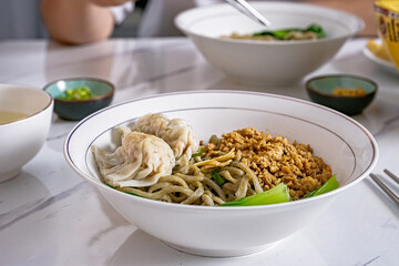 A white bowl of Asian noodles topped with minced pork, bok choy, and wontons on a marble table, with a person eating in the background, ideal for food photography