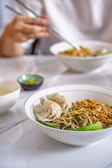 A white bowl of Asian noodles topped with minced pork, bok choy, and wontons on a marble table, with a person eating in the background, ideal for food photography
