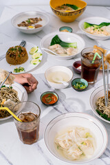 An overhead view of a shared meal with various Asian dishes, including noodles, rice, and soup, on a marble table, with hands using chopsticks and iced drinks, perfect for social dining 