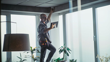 Male mature technician stands on ladder, installs CCTV camera in office room using tablet computer with professional software. Monitoring and tracking. Security system, video surveillance and privacy. © Framestock