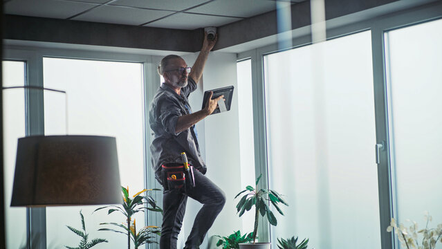 Male mature technician stands on ladder, installs CCTV camera in office room using tablet computer with professional software. Monitoring and tracking. Security system, video surveillance and privacy.