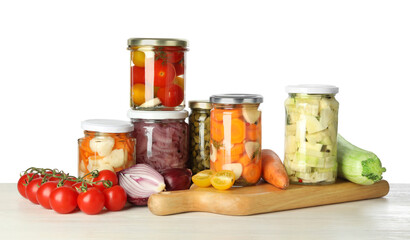 Different pickled products in jars and fresh vegetables on light wooden table against white background