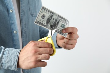 Budgeting. Man cutting dollar banknote with scissors on light grey background, closeup. Space for...