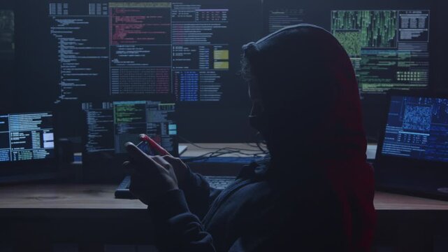 The distinctive silhouette of a hacker is visible while they are focused on multiple screens in a dark environment