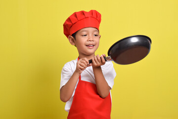 Cute little chef holding frying pan and spatula. Asian boy wearing red apron and hat on isolated...