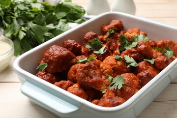 Baked cauliflower buffalo wings in baking dish and parsley on wooden table, closeup