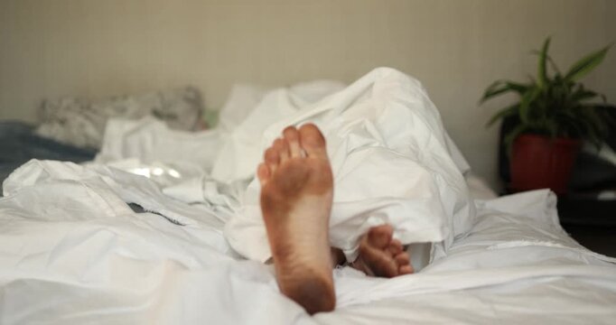 A cozy bedroom scene shows a man relaxing on a bed, his dirty and funny feet peeking out from under a soft white blanket, evoking calm and inviting a peaceful sleep.