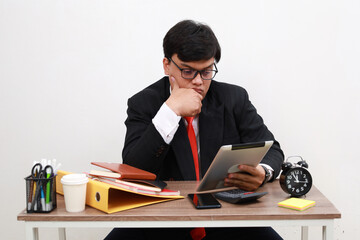 Unhappy male working in the office sitting on office desk while using tablet computer, front view, isolated on white.