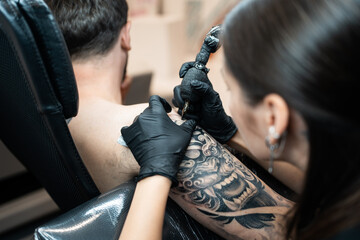 professional woman artist making stylish black tattoo of Japanese demon mask on man hand with machine and needle in salon, shoulder body art