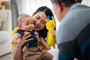 A man and a woman are playing with a baby in a living room. The baby is holding a toy and smiling. The scene is warm and friendly, with the family enjoying each other's company