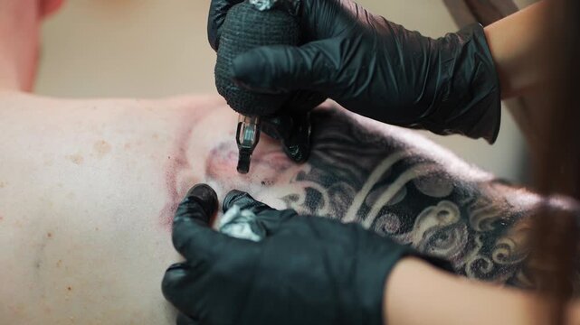 professional woman artist making stylish black tattoo of Japanese demon mask on man hand with machine and needle in salon, shoulder body art 4k