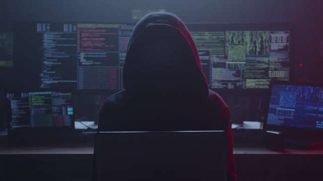 A shadowy silhouette of a hacker wearing a hoodie is surrounded by brightly glowing screens displaying various code