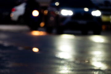 A blurry image of a car with its headlights on. The car is surrounded by other cars and the street is wet. Scene is dark and mysterious, with the blurred effect adding to the sense of movement