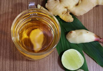 Ginger juice in a glass cup with lemon wedges, leaves and rhizomes on a dark wooden table.