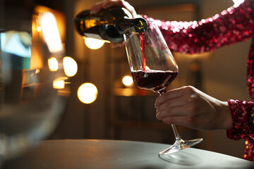 Woman pouring red wine from bottle into glass at table indoors, closeup