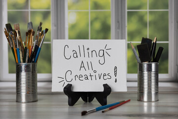 Calling all creatives! sign sitting on easel in front of a bay window with different sizes of paint...