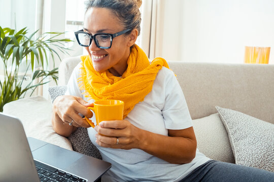 Front view of a happy adult woman working on her laptop during a video call, wearing a yellow scarf, smiling at the screen while enjoying a coffee or tea, reflecting online communication and social