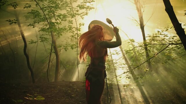 Halloween holiday concept. Dark silhouette fantasy woman witch dancing in forest ritual dance, fairy girl shaman playing tambourine, beating drum with stick melody magic spell. Fog sun rays light 4k