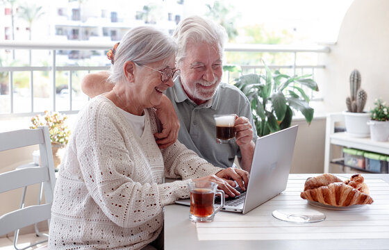 Smiling senior retired couple using together laptop while enjoying breakfast together sitting outside on home terrace with coffee, tea and baked goods. Peaceful retirement lifestyle.