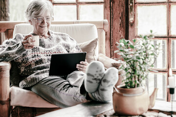Old 70s lady reading e-book on digital tablet at home, resting on armchair close the window in cozy...