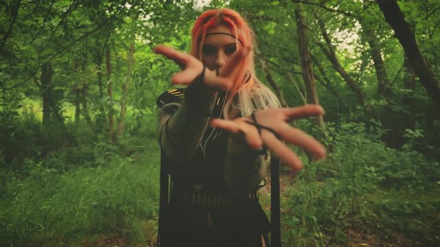 Halloween holiday woman witch conjuring with hands, dancing looking at camera walking in dark forest green tree. long black dress gothic boho style macrame, elf fairy girl shaman. orange colored hair.
