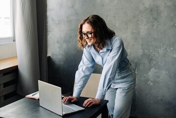 Professional woman with glasses sitting at a desk, working on a laptop in a bright office...