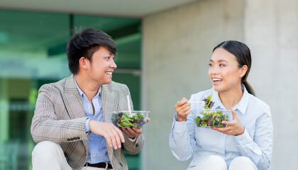Two young Asian adults man and woman are seated on a stone surface, sharing a light and healthy lunch consisting of fresh salads. They are smiling and engaged in a cheerful conversation