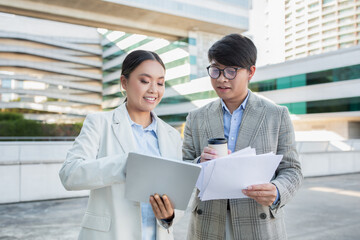 Two young Asian professionals are engaged in a collaborative discussion outside in an urban setting. The woman is holding a laptop and appears to be showing data