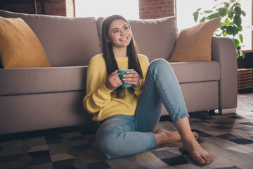 Full length photo of teen lady sit floor couch drink tea look empty space dressed yellow garment spend pastime spacious house indoors room