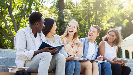 Excited multiracial group of students discussing new educational project, sitting with laptop and notepads on stairs in park