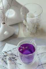 Purple lavender tea in transparent cup on plaid white towel, dried lavender flowers, vases and candle around on table, closeup lifestyle morning vertical still life in high key, idea of freshness