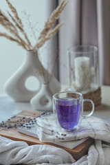Violet lavender tea in transparent cup on plaid white towel and wooden stand, bunch of dried lavender, vases and candle around, light lifestyle vertical still life, high key, idea of cozy morning