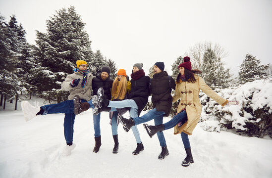 Friendly winter fun. Group of cheerful young people having fun dancing in snow while walking in winter forest. Group of friends imitates cancan dance standing in row with raised legs in snowy area.