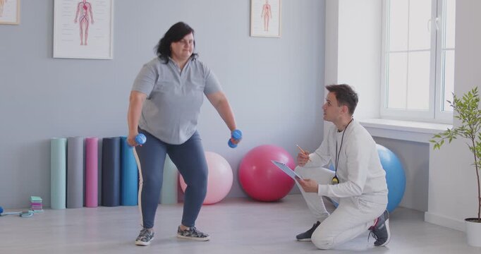 A physiotherapist helps a young plump woman squat with dumbbells. A rehabilitation specialist trains a patient after an injury. Concept active rehabilitation, medical rehabilitation center