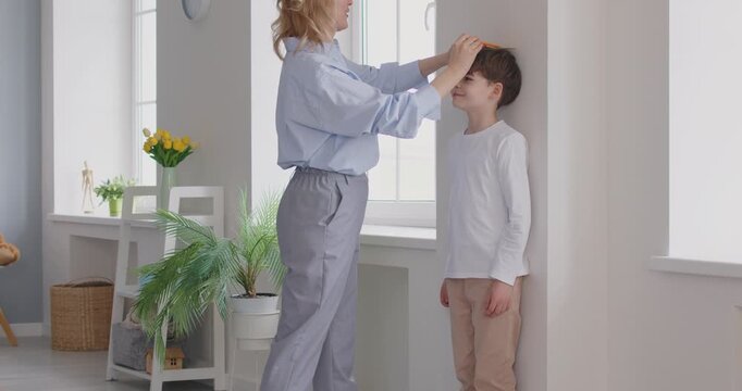 Mom uses a ruler and a pencil to measure and mark the height of her child. Happy, loving mother gives a high five to her son after measuring his height on a wall at home. Growing up concept