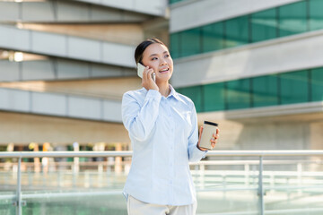 Fototapeta premium A professional Asian woman dressed in a light blue shirt stands outdoors, smiling as she engages in a phone conversation. She holds a takeaway coffee cup in her left hand, copy space