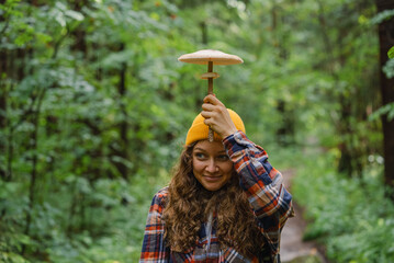 Woman wearing yellow beanie holds large mushroom above her head like an umbrella while during the rain in forest. 