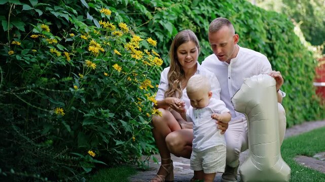 Loving mom and dad sit squatted near their little son. Cute blond baby boy drops the flower and picks it up.